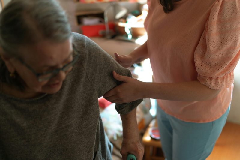 A caregiver helps an elderly woman stand up, gently holding her arm for support.  Image courtesy of Pexels.
