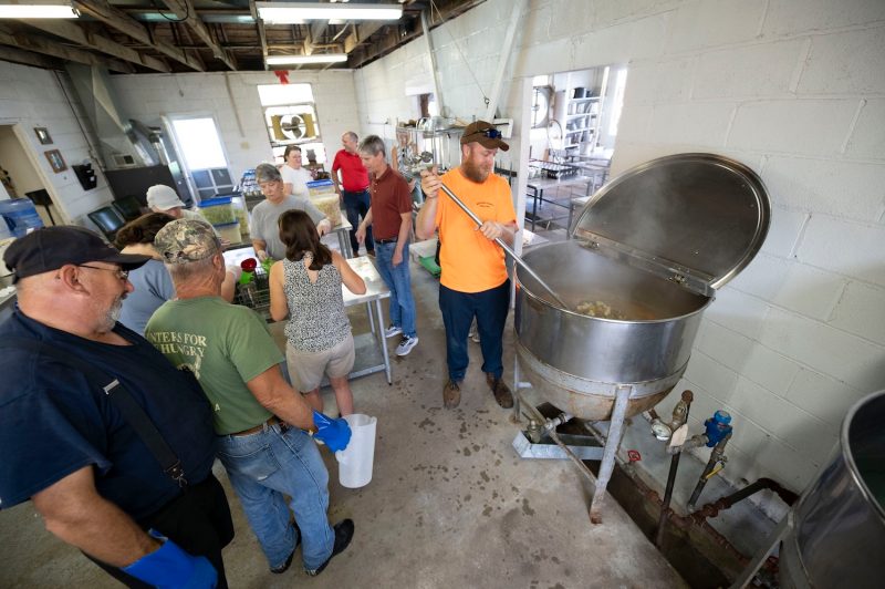 People stand in a large industrial kitchen as a man stirs a pot.