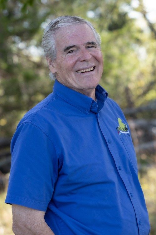 Headshot of a man with grey hair and a blue shirt with logo, smiling broadly at the camera.