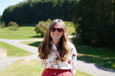 Woman wears sunglasses outside while smiling at the camera.