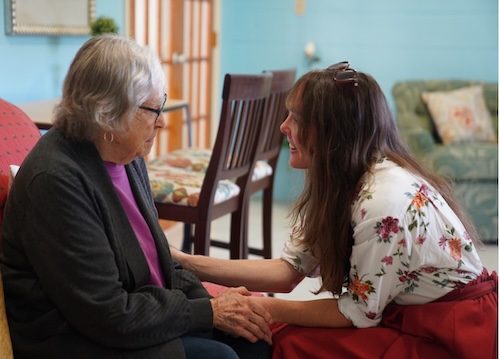 A woman with grey hair sits in a chair and looks down at a younger woman who crouches in front of her and smiles.