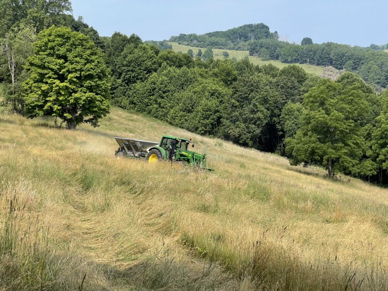 A green tractor with yellow wheels operating in a grassy field, surrounded by dense trees and rolling hills in the background.