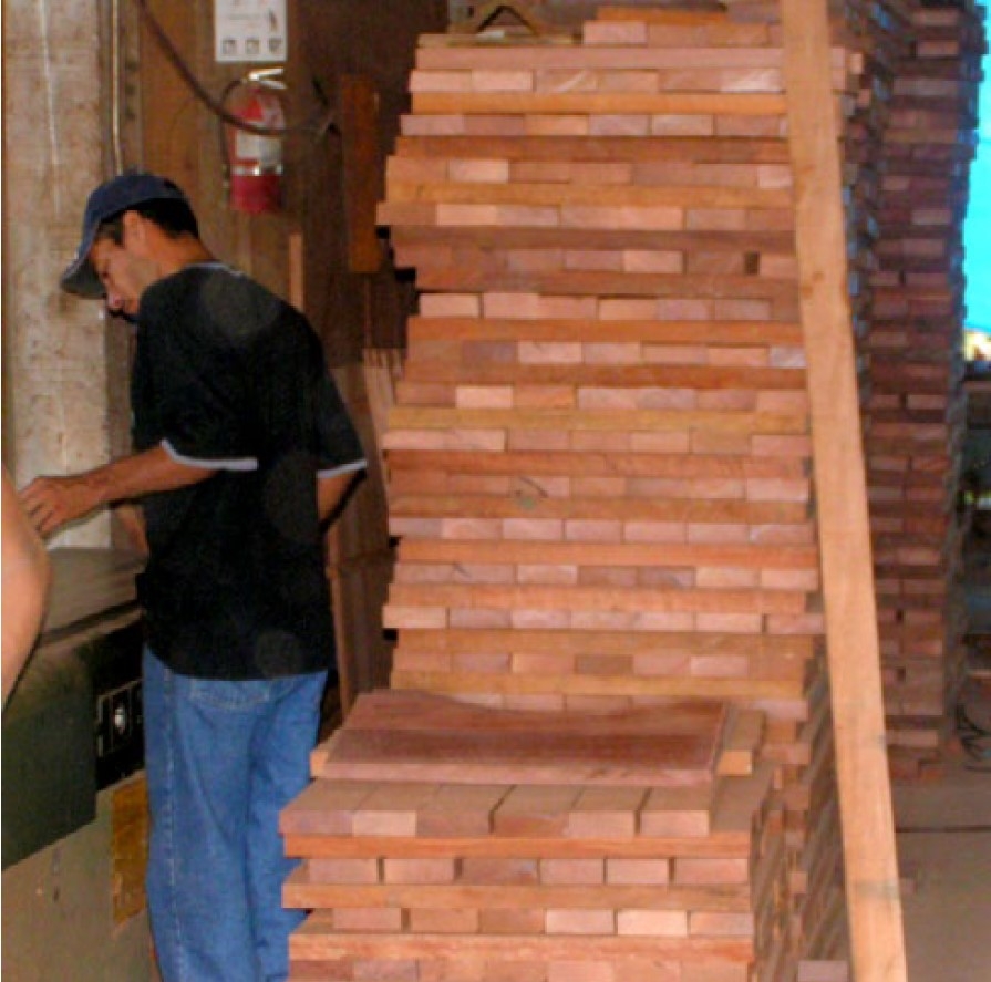 A person in a black shirt and blue jeans standing in front of piles of seemingly wood planks.