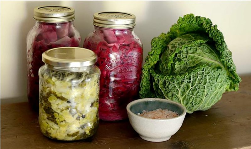 Canning jars of pickled sauerkraut, a head of green cabbage, and a small bowl of canning salt.