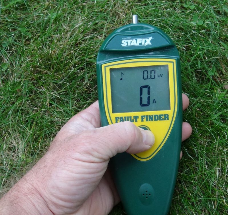 A hand holding a Stafix brand fault finder over a grassy area; the screen shows 0.0 kilovolts and 0 amps.